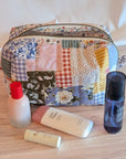 A patchwork fabric cosmetic pouch with a curved top and zip closure, displayed on a gray wood floor next to a potted green plant against a white paneled wall.