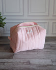 A light pink quilted cosmetic bag with a small white bow detail, sitting on a gray wood floor in front of a white paneled wall with a potted plant on the left.