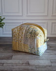 A quilted fabric zippered pouch in a yellow and floral patchwork pattern, sitting on a gray wood floor in front of a white paneled wall with a potted plant on the left.