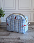 A light blue and white gingham toiletry bag with small red embroidered cherries, sitting on a gray wood floor in front of a white paneled wall.