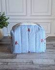 A light blue and white gingham toiletry bag with red cherries and green leaves embroidered on the front, sitting on a gray wood floor in front of a white paneled wall.