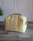 A yellow and white gingham toiletry bag with a small white bow on the front, sitting on a gray wood floor in front of a white paneled wall next to a potted green plant.