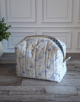 A quilted cosmetic pouch with a blue floral pattern on a cream background, sitting on a gray wood floor in front of a white paneled wall.