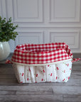 A quilted white cosmetic bag with a red cherry print and red gingham trim, sitting on a gray wood floor next to a potted green plant.