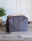 A quilted cosmetic pouch with a navy blue base and a small pink, orange, and yellow floral pattern, sitting on a gray wood floor in front of a white paneled wall with a potted plant on the left.