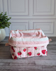 A quilted cosmetic pouch with a pink and white strawberry pattern, sitting on a gray wooden floor in front of a white paneled wall with a potted plant on the left.