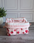 A quilted cosmetic pouch with a pink and white strawberry pattern, sitting on a gray wooden floor in front of a white paneled wall with a potted plant on the left.