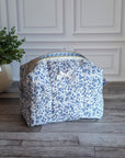 A fabric cosmetic bag with a blue and white floral print sits on a gray wood plank floor in front of a white paneled wall, next to a potted green plant.