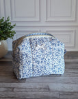 A fabric cosmetic bag with a blue and white floral print sits on a gray wood plank floor in front of a white paneled wall, next to a potted green plant.