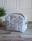 A fabric cosmetic bag with a blue and white floral print sits on a gray wood plank floor in front of a white paneled wall, next to a potted green plant.