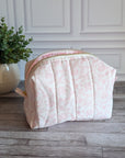 A light pink and white patterned fabric cosmetic pouch with a pleated design, sitting on a gray wood floor next to a potted green plant against a white paneled wall.