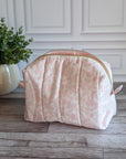 A light pink floral print zippered cosmetic pouch or case sits on a gray wood floor in front of a white paneled wall, with a potted green plant to the left.