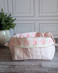 A pink and white checkered lining fabric quilted pouch, placed on a gray wood floor in front of a white paneled wall, with a potted green plant on the left.