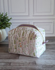 A quilted cosmetic bag with a pink and green floral print sits on a gray wood floor in front of a white paneled wall, next to a potted green plant.