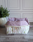 A quilted cosmetic bag with a pink and green floral print sits on a gray wood floor in front of a white paneled wall, next to a potted green plant.