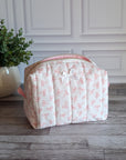 A quilted cosmetic bag with a pink and white bow pattern, sitting on a gray wood floor in front of a white paneled wall next to a potted green plant.