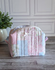 A patchwork quilted cosmetic bag in pastel pink, blue, and white fabrics, sitting on a gray wooden floor in front of a white paneled wall with a potted plant on the left.