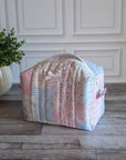 A patchwork quilted cosmetic bag in pastel pink, blue, and white fabrics, sitting on a gray wooden floor in front of a white paneled wall with a potted plant on the left.