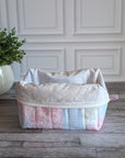 A patchwork quilted cosmetic bag in pastel pink, blue, and white fabrics, sitting on a gray wooden floor in front of a white paneled wall with a potted plant on the left.