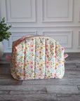 A quilted cosmetic pouch with a pink and yellow floral pattern on a white background, sitting on a gray wood floor next to a potted green plant.