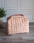 A quilted cosmetic pouch in a light peach-pink color with a pattern of small orange and pink strawberries, standing on a gray wood floor next to a potted green plant.