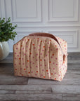 A quilted cosmetic pouch in a light peach-pink color with a pattern of small orange and pink strawberries, standing on a gray wood floor next to a potted green plant.