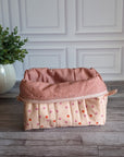 A quilted cosmetic pouch in a light peach-pink color with a pattern of small orange and pink strawberries, standing on a gray wood floor next to a potted green plant.