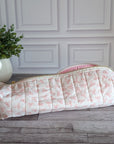 A quilted fabric zippered pouch with a pink bow pattern on a cream background, resting on a gray wood floor in front of a white paneled wall with a potted green plant.