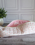 A quilted cosmetic pouch with a pink and white floral print and a pink gingham lining, resting on a gray wooden floor in front of a white paneled wall.