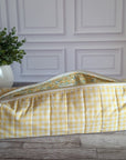 A yellow and white gingham quilted travel or toiletry bag with a floral patterned lining, resting on a rustic wooden floor in front of a white paneled wall.