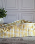 A yellow and white gingham quilted travel or toiletry bag with a floral patterned lining, resting on a rustic wooden floor in front of a white paneled wall.