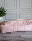 A light pink quilted fabric zippered pouch or cosmetic bag resting on a gray wood floor in front of a white paneled wall, with a potted green plant on the left.