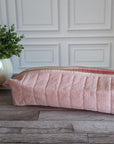 A light pink textured zippered pouch or cosmetic bag resting on a rustic wooden floor in front of a white paneled wall, with a potted green plant on the left.