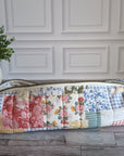 A long rectangular quilted patchwork zippered pouch or case with a floral and gingham pattern, resting on a gray wood floor in front of a white paneled wall with a potted green plant on the left.