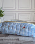A light blue and white gingham zippered pouch with embroidered red cherries, resting on a gray wood floor next to a potted green plant.