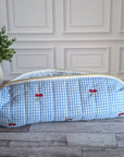 A light blue and white gingham zippered pouch with small red cherry prints, resting on a gray wooden floor in front of a white paneled wall.