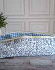 A quilted fabric storage bag with a blue and white floral print exterior and a blue and white gingham lining, resting on a gray wood floor in front of a white paneled wall.