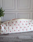 A quilted white fabric bag with a red cherry print pattern, resting on a gray wooden floor in front of a white paneled wall with a potted green plant on the left.