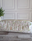 A quilted cylindrical cosmetic pouch with a delicate pink and green floral print, resting on a gray wood floor next to a potted green plant against a white paneled wall.