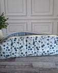 A quilted fabric zippered pouch with a blue and white floral print and a blue-and-white gingham lining, resting on a gray wood floor next to a potted green plant.