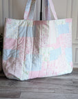 A quilted patchwork tote bag in pastel pink, blue, and white fabrics, standing on a gray wood floor next to a potted green plant.