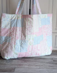 A large quilted patchwork tote bag in pastel pink, blue, and white fabrics, standing on a gray wood floor against a white paneled wall with a potted plant on the left.
