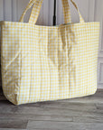 A large quilted tote bag with a yellow and white gingham check pattern, standing on a gray wood floor next to a potted green plant.