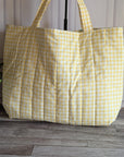A large quilted tote bag in a yellow and white gingham check pattern, standing on a gray wood floor against a white paneled wall with a potted green plant on the left.