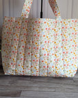 A quilted fabric tote bag with a small multicolor floral print, standing on a gray wood floor next to a potted green plant.