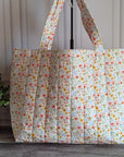 A large quilted tote bag with a small multicolor floral print on a cream background, standing on a gray wood floor next to a potted green plant.