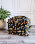 A black zippered cosmetic pouch decorated with a colorful butterfly pattern, set on a gray wood floor next to a potted green plant against a white paneled wall.