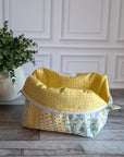 A handmade patchwork cosmetic bag made from yellow, white, and floral fabrics, sitting on a gray wood floor in front of a white paneled wall with a potted plant on the left.