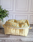 A fabric cosmetic pouch with a yellow and white gingham outer and a yellow floral top lining, placed on a rustic wood floor next to a potted green plant against a white paneled wall.