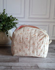 A quilted cosmetic pouch with a cream background and a repeating cherry-blossom pattern, set on a gray wood floor next to a potted green plant against a white paneled wall.